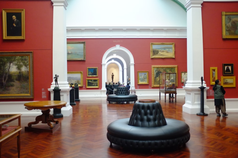 A spacious art gallery with red walls, wooden floors, black circular seating, and various framed paintings and sculptures on display. Several people observe the artworks. Natural light streams in from above.