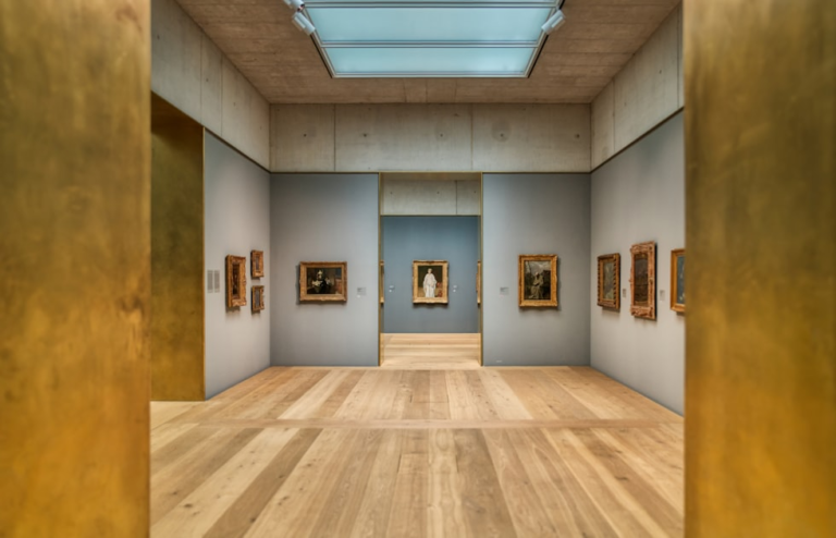 A modern art gallery with gold-colored door frames, wooden floors, gray walls, and framed paintings displayed in well-lit, spacious rooms under a large skylight.