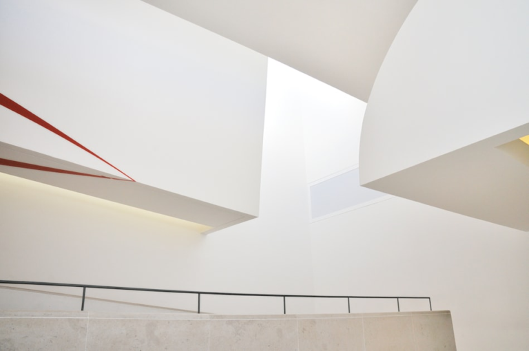 Interior view of a modern, minimalist building with white walls, geometric shapes, soft natural lighting, and a single thin red diagonal line as an accent. A simple railing follows a sloped walkway below.