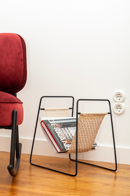 A modern magazine holder with a woven sling holds several magazines next to a red upholstered chair on a wooden floor against a white wall with two power outlets.