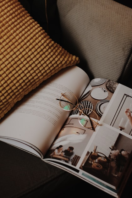 An open magazine with photos and text lies on a couch beside a mustard-yellow textured pillow, with a pair of eyeglasses resting on the pages.