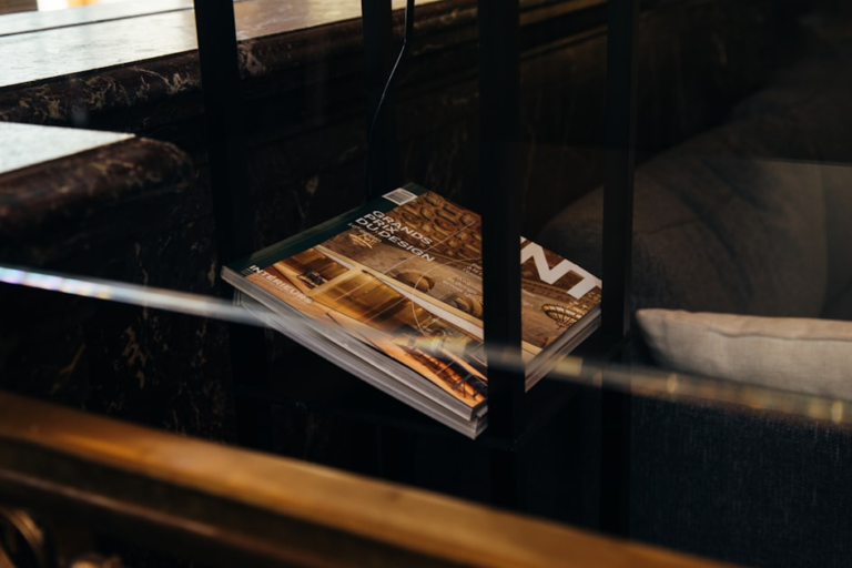 A stack of magazines with architectural and interior design covers sits on a black metal shelf next to a marble counter and a gray cushion.