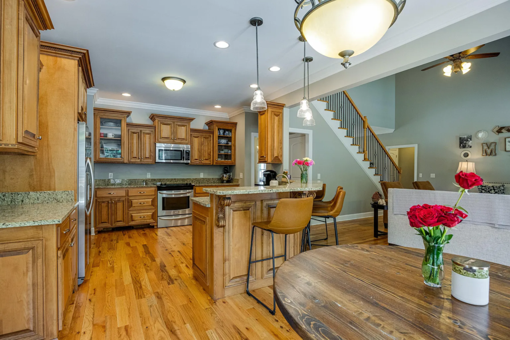 Experience a stunning kitchen transformation featuring wood cabinets, stainless steel appliances, and an island with two tan bar stools. The nearby dining table with red roses and the living area with a staircase complete the inviting space.