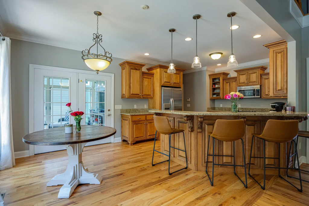 Experience a kitchen transformation with wooden cabinets, granite countertops, and an island with three brown barstools. A round dining table with flowers sits nearby, illuminated by pendant lights and natural light from French doors.