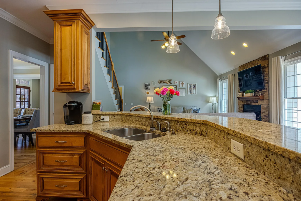 Experience a stunning kitchen transformation with granite countertops, wooden cabinets, a sink, and pendant lights, seamlessly flowing into a living room featuring a gray sofa, wall art, TV over the fireplace, and a vase of flowers on the counter.
