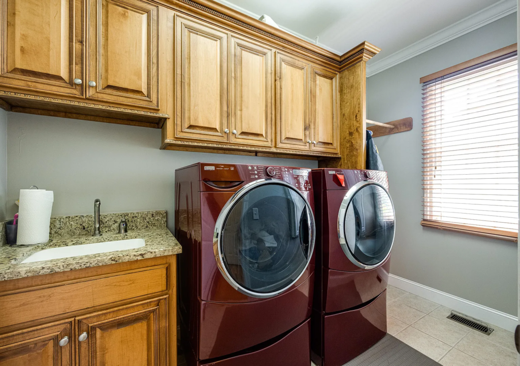 A laundry room with red front-loading washer and dryer, wooden cabinets, granite countertop with a sink, paper towels, and a window with blinds letting in natural light—perfect for your next kitchen transformation.